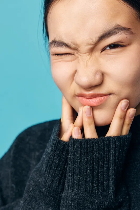 Woman with natural skin, wearing black sweater, touching her face with fingers, eyes closed and expression of discomfort on blue background. Closeup portrait of person with hand near chin.