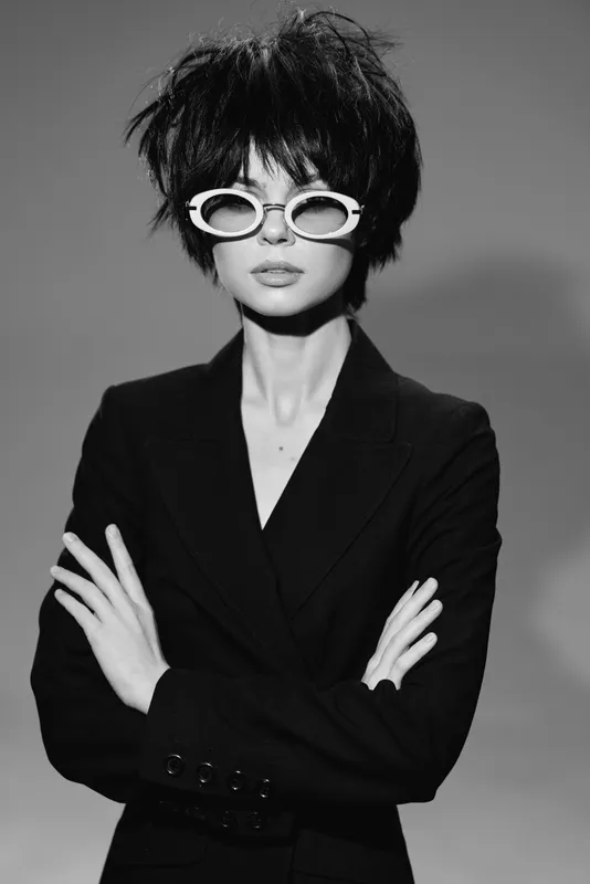 Black and white portrait of a stylish woman wearing white sunglasses and a dark suit with arms crossed.