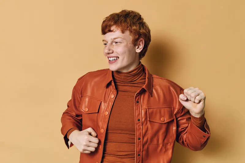Young man with red hair wearing orange jacket and turtleneck smiling and dancing against beige background for happiness and casual style.