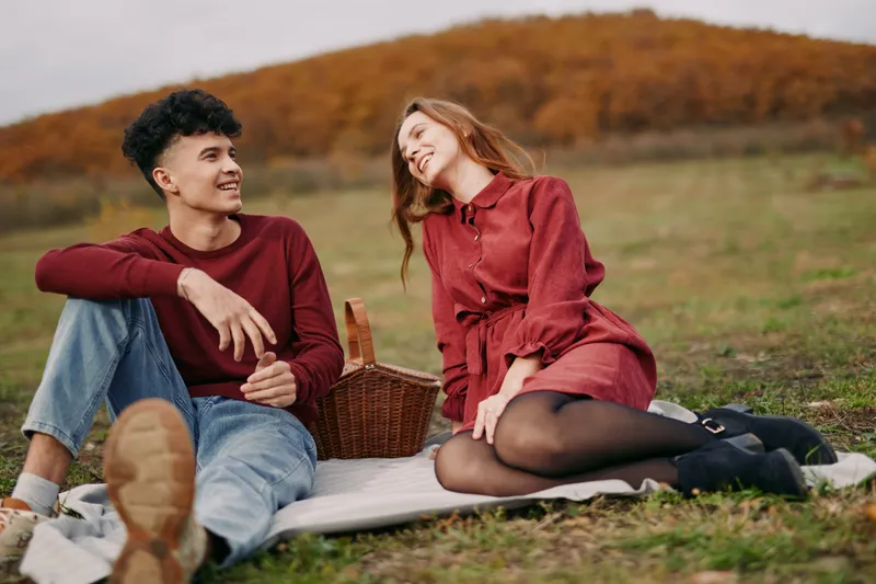 Two friends share a candid autumn picnic on a sunlit field, with genuine smiles, relaxed poses, and spontaneous interaction that convey authenticity.