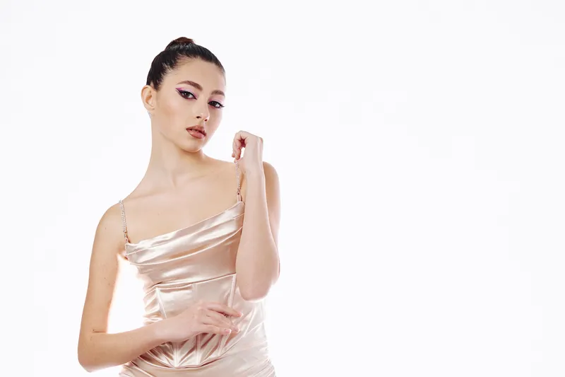 Elegant young woman in a silky beige dress poses with a thoughtful expression and arm bent. Soft makeup highlights her eyes and lips. Studio shot on a white background. Beauty fashion concept.