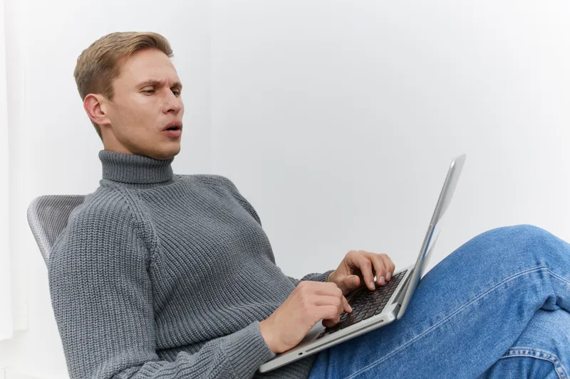 Relaxed young man with fair hair wearing gray turtleneck sweater using silver laptop sitting on a white background, casual indoor scene, leisure activity, technology, modern lifestyle.