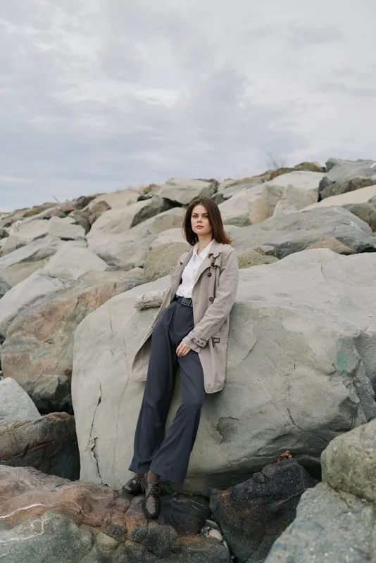 Young woman in casual outfit stands on rocky coast under cloudy sky. Natural pose with neutral expression captures serene outdoor lifestyle and fashion concept.