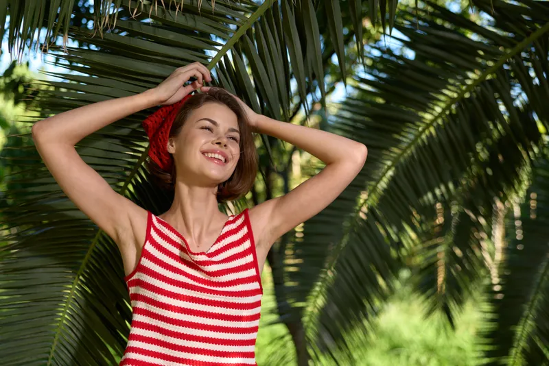 A cheerful woman wearing a red and white striped top stands among lush tropical palm leaves, smiling warmly as bright sunlight highlights her relaxed, carefree mood in an outdoor setting.