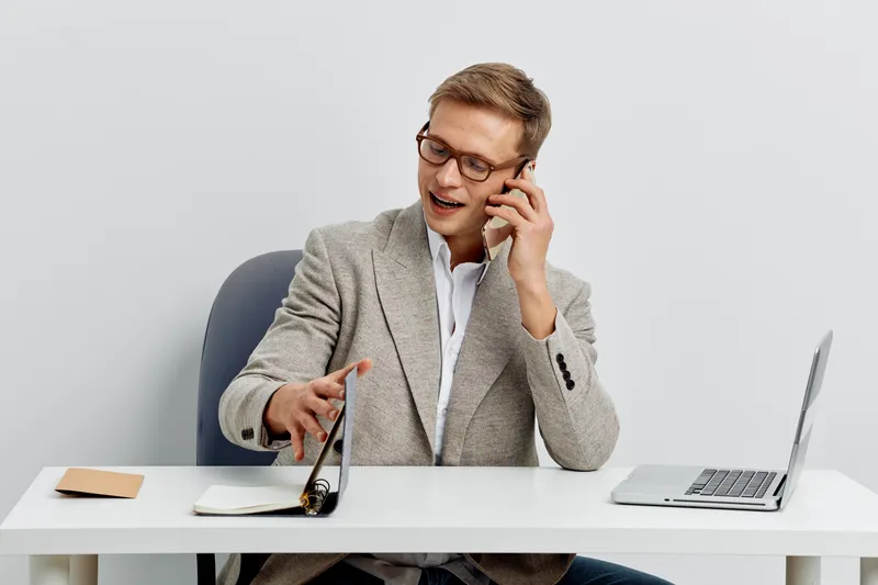 Professional middle-aged smiling man talking on mobile phone while working at modern office desk with laptop and notebook, dressed in smart casual wear. People business concept.