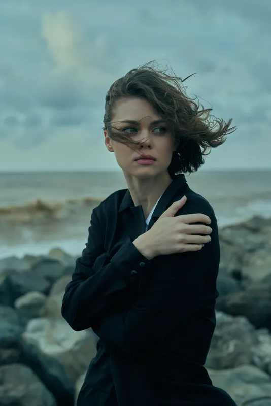 Woman in a dark coat stands by a rocky shoreline, wind sweeps hair, moody ocean backdrop, cinematic composition emphasizes resilience and mystery, stormy atmosphere, rugged coast