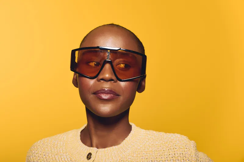 Portrait of confident black woman with dark skin wearing oversized orange sunglasses and beige knit sweater on yellow background. Concept of fashion, style, and modern accessories.