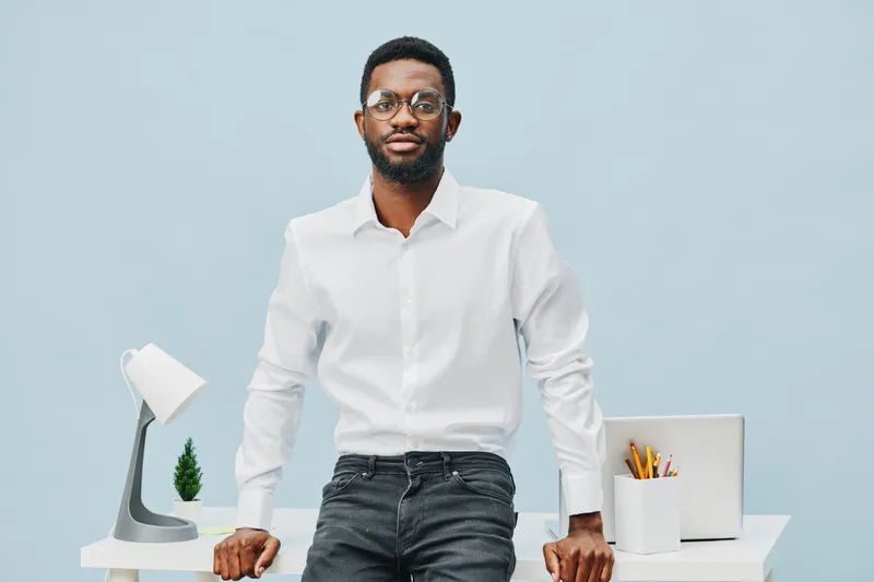 Confident young African American man in white shirt standing behind office desk with modern lamp, potted plant, and stationery, indoors. Business, professional, workspace, portrait, modern office envi
