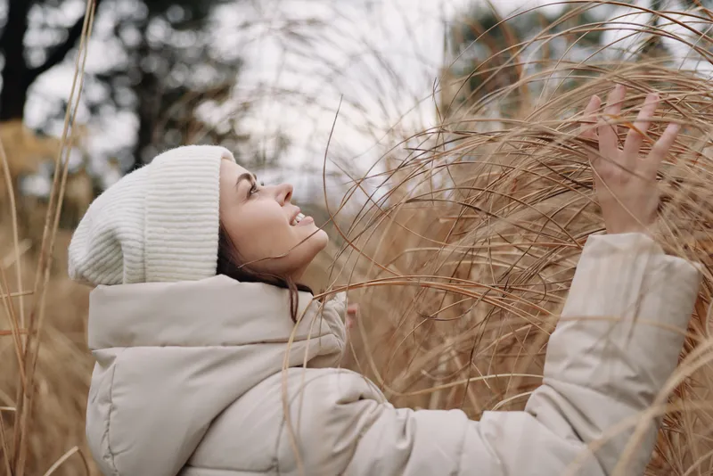 Winter scene with a woman in a puffy coat and knit beanie reaching through tall grass, capturing a calm outdoor moment among rustic reeds.