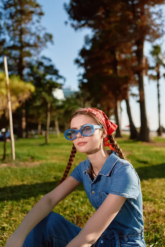 lifestyle woman in modern boho-western denim wearing blue sunglasses and red bandana sitting on grass outdoors with warm film color tones and relaxed street style vibe