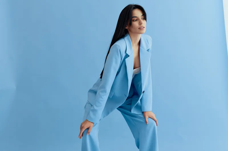 young woman in blue suit posing confidently against a light blue background, showcasing modern fashion with long dark hair and a relaxed posture