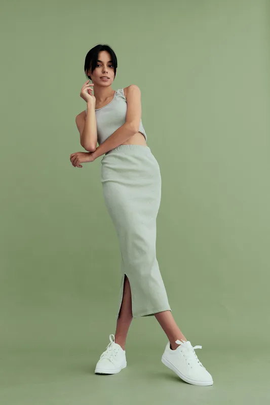 Stylish woman wearing a light green matching top and skirt with white sneakers posing confidently against a plain green background in a minimalistic studio setting.