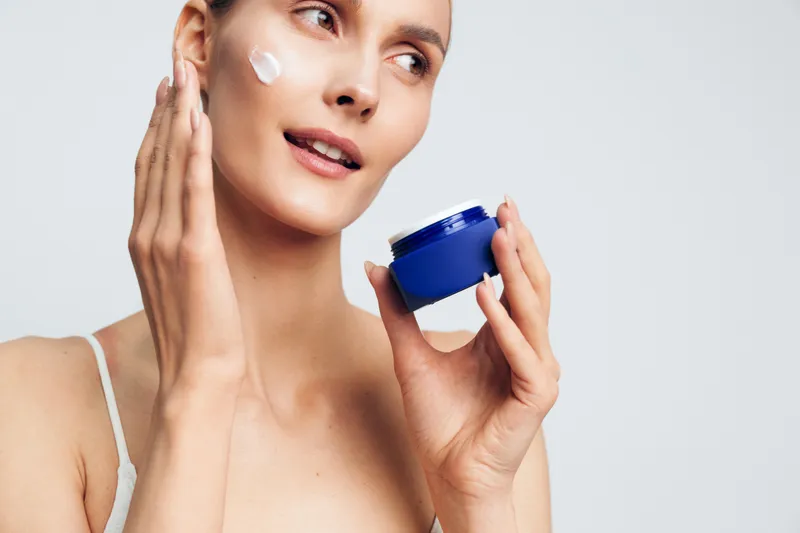 Young woman with clear skin applies cream to her face while holding a blue jar in a bright studio, showcasing skincare routine and natural beauty care.