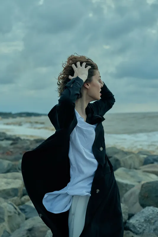 Windy seashore scene with a windblown coat, a woman among rocks, sea and ocean behind, motion in her stance as gusts sweep the shore under a moody sky
