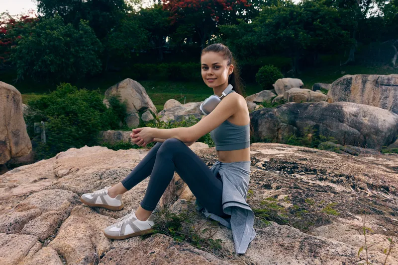 Young woman in sportswear sitting on rocky terrain, enjoying nature, conveying a sense of fitness, relaxation, and outdoor activity amidst greenery