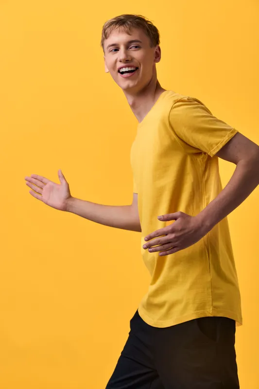 Joyful man in a yellow shirt moves across a bright studio background, expressive smile, light steps and dynamic pose conveying energy, optimism and movement in a cheerful fashion shoot