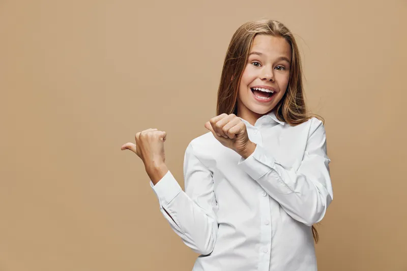 Girl with long hair and white shirt smiling and pointing with fingers to side on beige background. Child portrait with positive expression and casual look in studio.