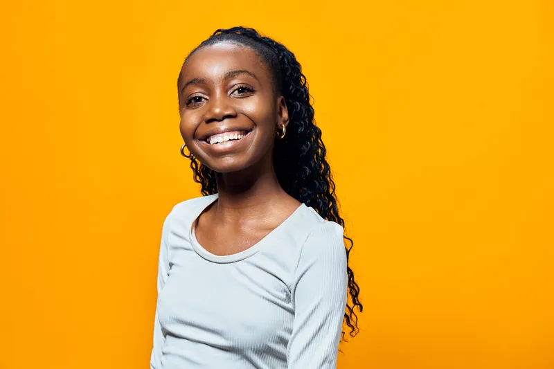 Smiling young Black girl with curly hair, wearing a light grey shirt against a vibrant yellow background, expressing joy and confidence in a playful pose