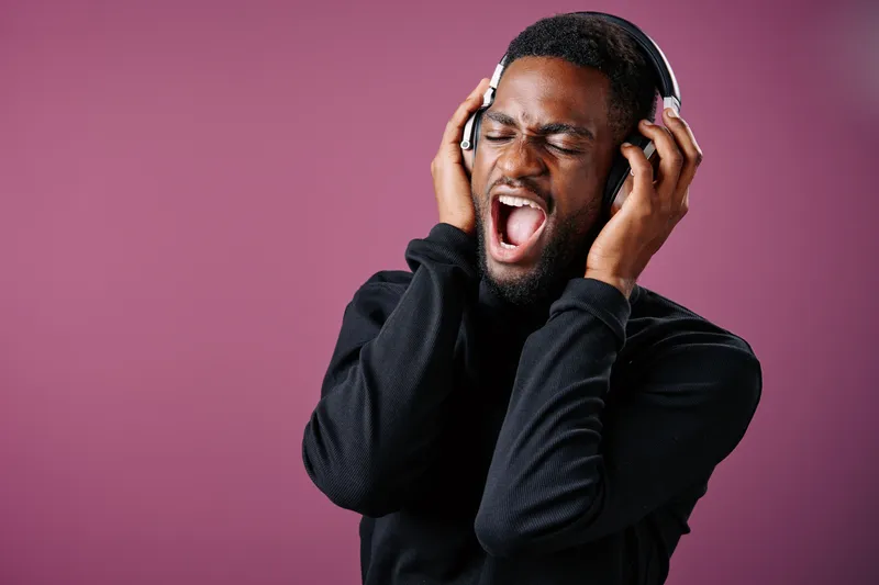 Young Black man passionately singing while wearing headphones, expressing joy and energy against a vibrant pink background