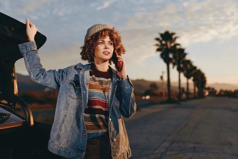 Woman lifestyle sunset car denim jacket road outdoor, curly haired woman stands beside car on evening street, striped sweater under denim jacket, relaxed travel pose, warm golden light