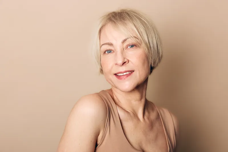 Smiling middle aged woman with short blonde hair wears beige tank top in studio on neutral background. Concept of natural beauty, confidence, aging gracefully, wellness, and positive mood.