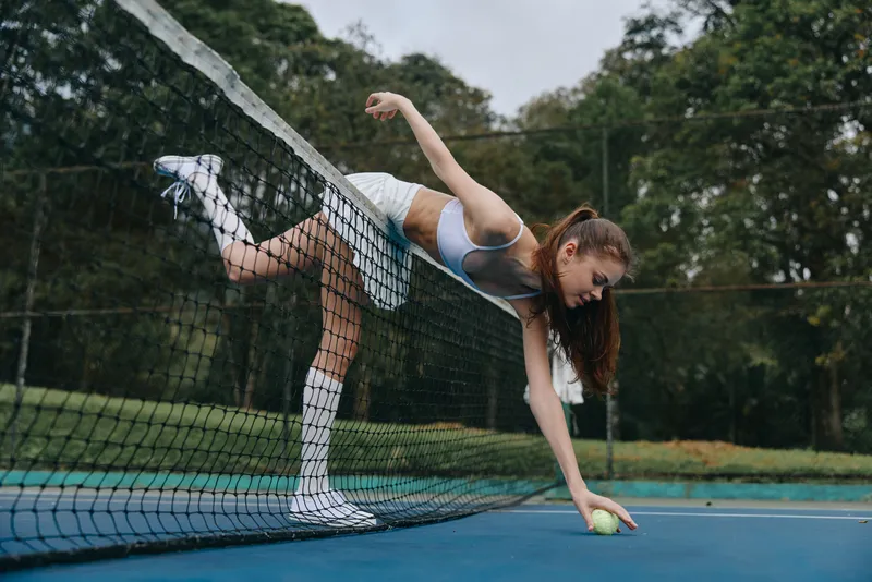 Young athletic woman in sportswear reaches for a tennis ball on a court, showcasing her dedication and passion for sports, surrounded by lush greenery and a vibrant atmosphere