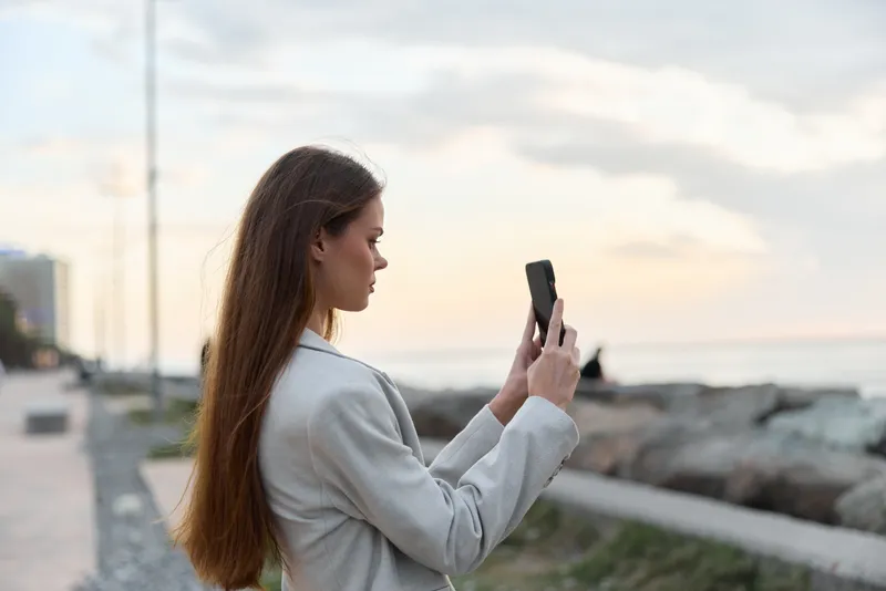 Young woman with long brown hair taking photo on smartphone near rocky sea shore at sunset, casual style, outdoor lifestyle concept