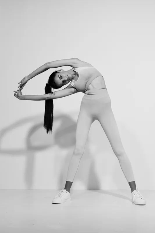 Black and white woman sport fitness stretching sideways in stylish sportswear with long dark hair, demonstrating flexibility and strength against a plain studio background.