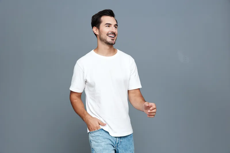 Smiling cheerful handsome young man in casual white t-shirt walks happily against plain gray background. Lifestyle portrait showcasing joy and confidence.