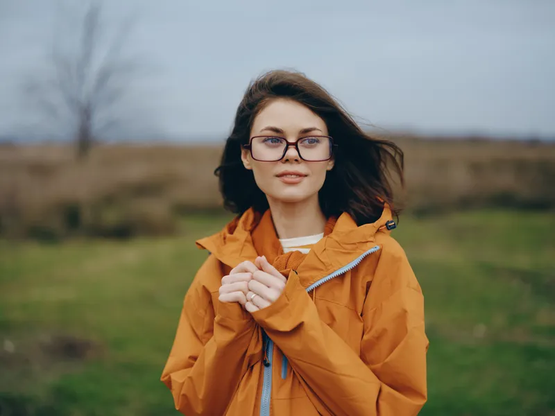 Smiling woman outdoor lifestyle portrait with autumn jacket and glasses, wind moving hair. Countryside field background, rural landscape, soft overcast daylight, relaxed travel mood today.