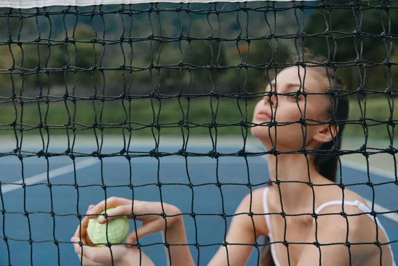 Young woman in sports attire holding a tennis ball, captured behind the net on an outdoor court, conveying focus and determination during a sunny day