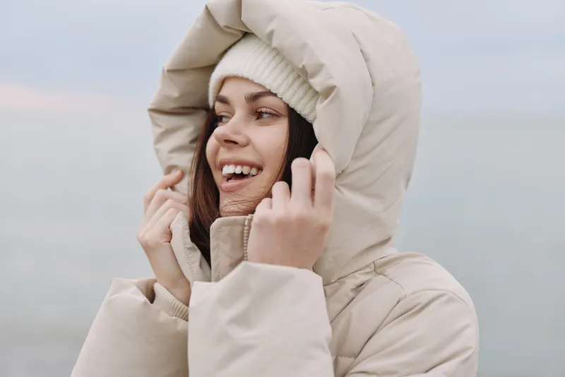 Woman in a warm beige hooded coat smiles by the seaside, cool breeze and outdoor winter portrait, casual chic fashion and relaxed mood. Night mood.