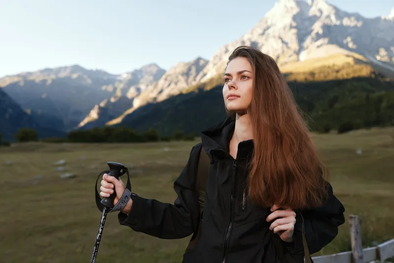 A lone female traveler stands in a scenic mountain landscape, gripping a trekking pole and gazing into the distance with calm resolve.