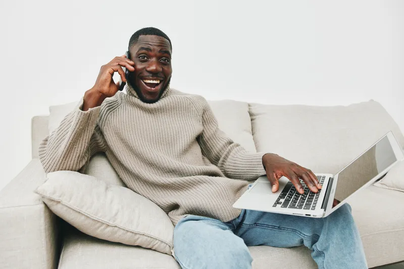 Happy African American man talking on the phone while working on a laptop, wearing a cozy sweater, sitting on a light sofa in a minimalist room