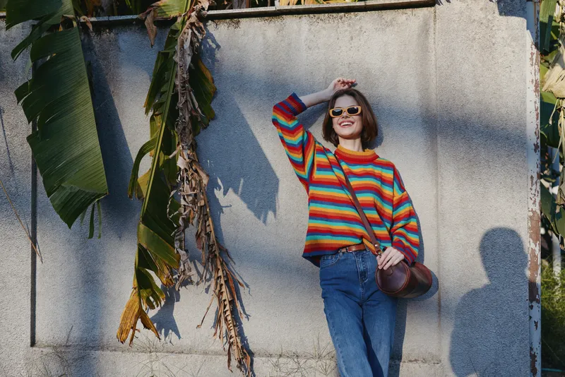 Woman in her mid 20s wearing a colorful striped sweater and sunglasses, smiling while leaning against a concrete wall with tropical outdoor banana leaves, casual outfit travel vibe.