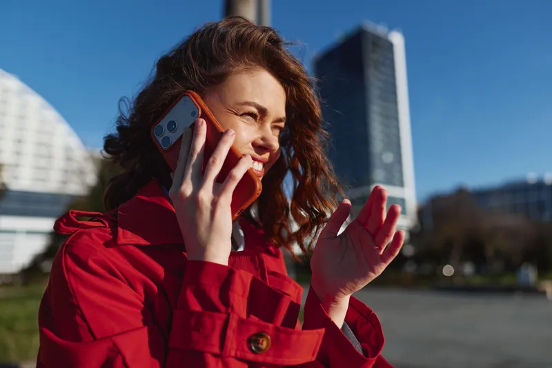 Young woman with curly hair wearing a red coat talks on a smartphone outdoors in a modern urban area on a sunny day with a bright blue sky.