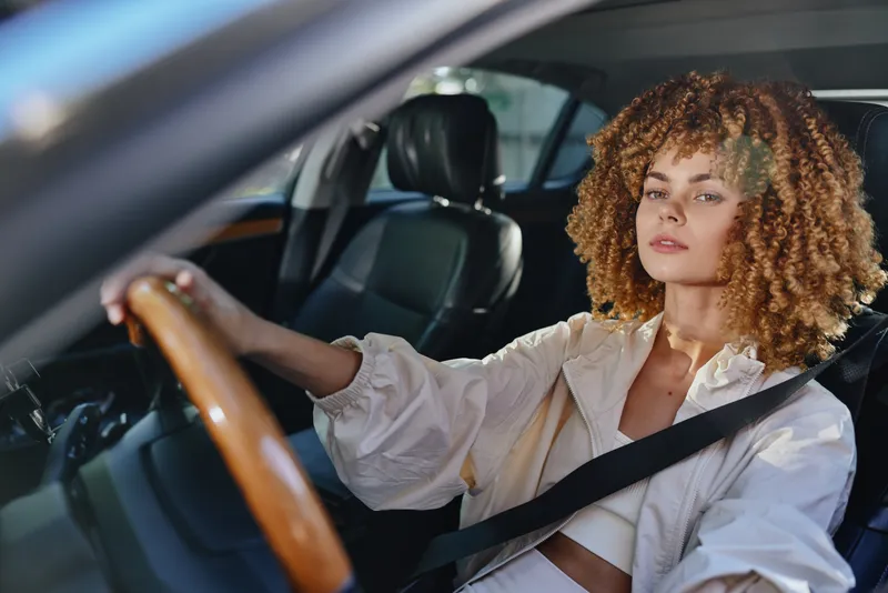 young woman driving car wearing seatbelt with curly hair and casual jacket, focused expression, daylight, modern vehicle interior, safe driving concept