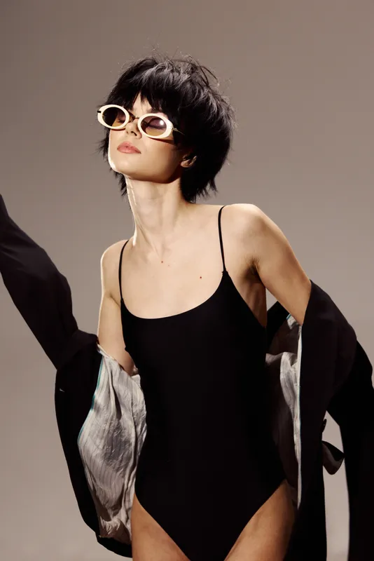 Stylish woman in black swimsuit wearing sunglasses and black jacket with confident expression in studio setting.
