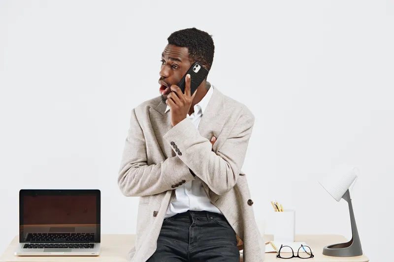 Businessman with headphones talking on mobile phone sits at desk with laptop and office supplies in bright modern workspace, concept of communication, technology, remote work, office environment, mult