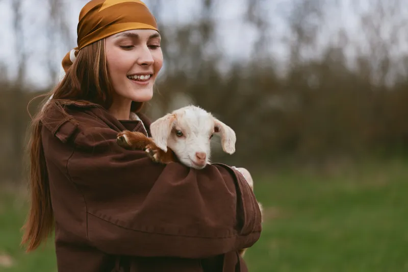 Cheerful woman outdoors, embracing a young goat kid in a sunlit field, relaxed pose, warm earth tones, rustic clothing, gentle connection, natural colors, tranquil countryside backdrop, candid