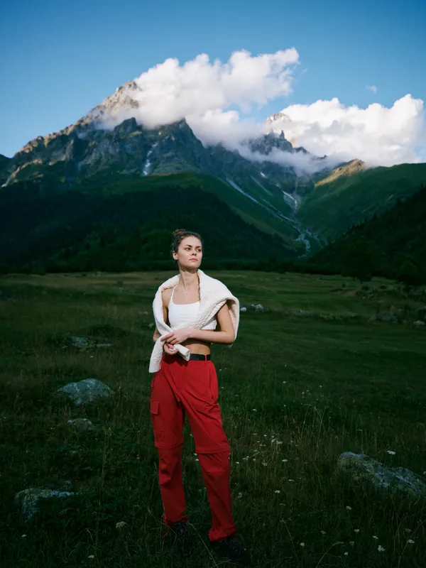 Woman standing in a grassy meadow with red pants and a draped sweater, alpine mountains and cloudy blue sky behind, capturing outdoor exploration, nature, and expansive landscape