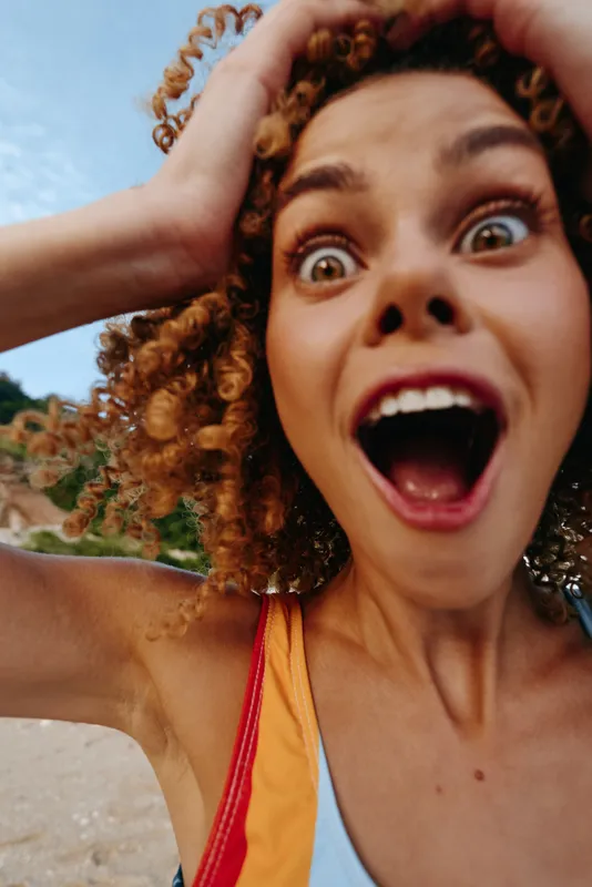 Woman curly hair surprise outdoors summer sunlight portrait, open mouth joyful expression, hands holding hair by ears, close up selfie outdoors, warm breeze, natural colors, beach background