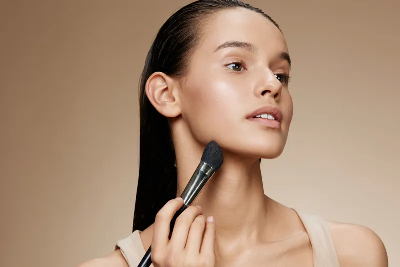 Young woman applying makeup with a brush, showcasing natural beauty and confidence in a soft, neutral background Perfect for beauty, skincare, and cosmetic themes