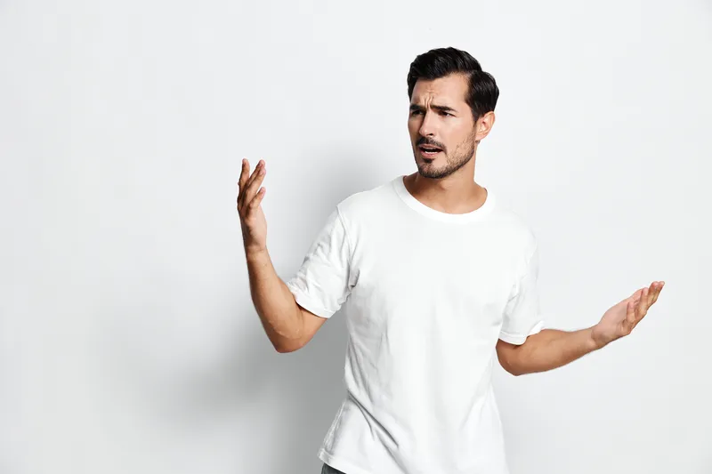 Confused young man in casual white t-shirt looking at camera with hands raised, expressing uncertainty isolated on plain solid light background. People emotion concept.