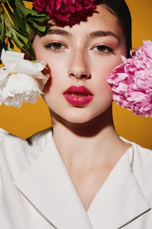 Close-up of a woman with glossy dark red lips and natural makeup surrounded by vibrant pink and white flowers. Stylish beauty portrait on bright yellow background of fashion.