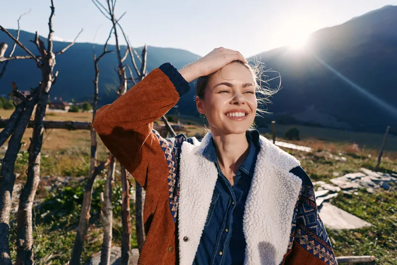 Woman with a bright smile standing in front of mountains under the sun outdoors, wearing a warm jacket in the countryside, happy portrait conveying joy, sunlight and rustic leisure.
