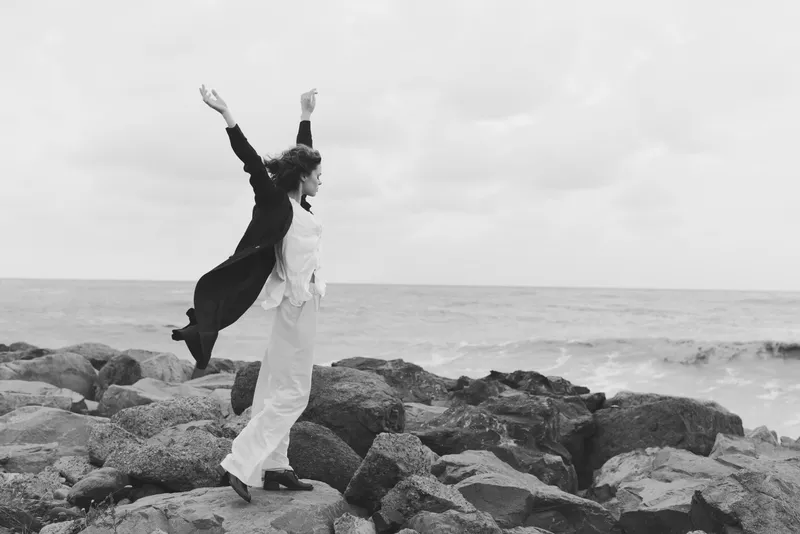 Woman leaping on rocky shore by the sea, celebration and freedom in motion as wind lifts a coat, black and white scene capturing bold exploration and coastal joy.