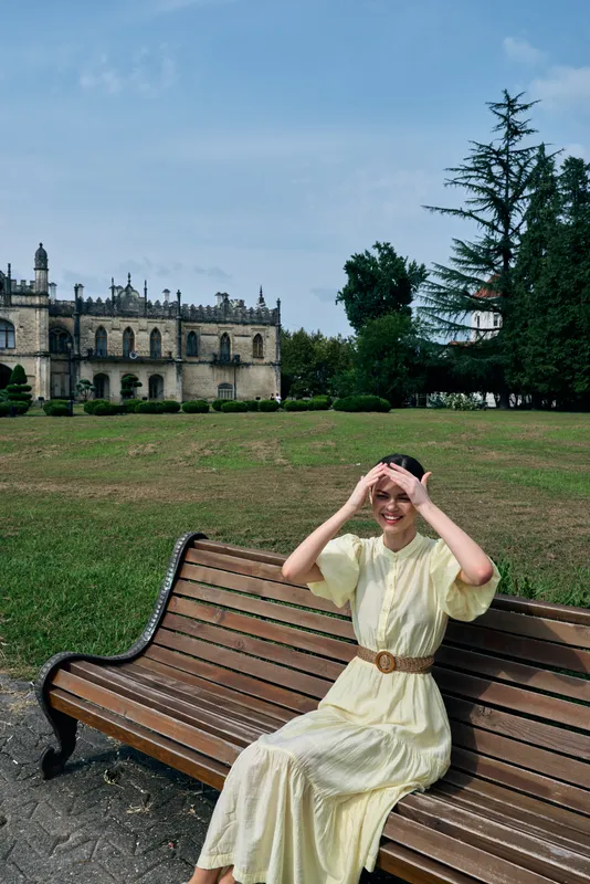 Woman on bench in park wearing vintage yellow dress smiling and making binocular hand gesture authentic inclusive portrait near manor