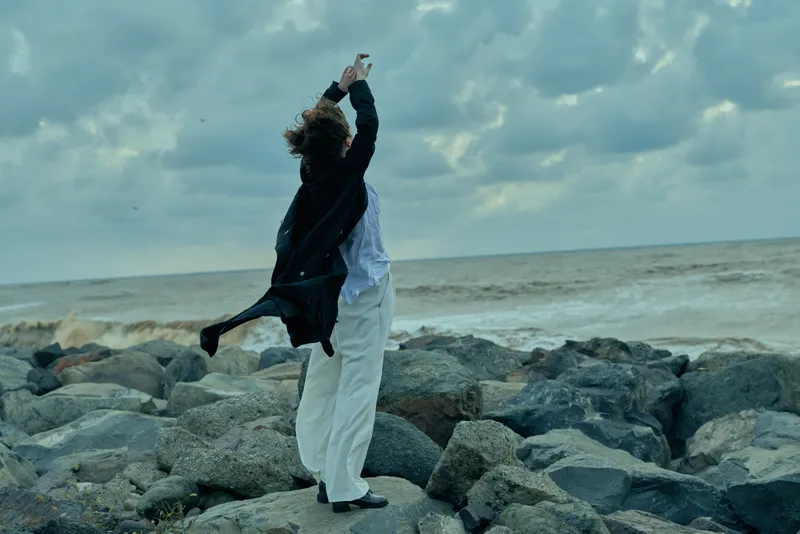 Two people on a rocky coastline share a joyful moment as they dance, arms raised toward a cloudy sky, with salt air and crashing waves framing a scene of freedom and connection