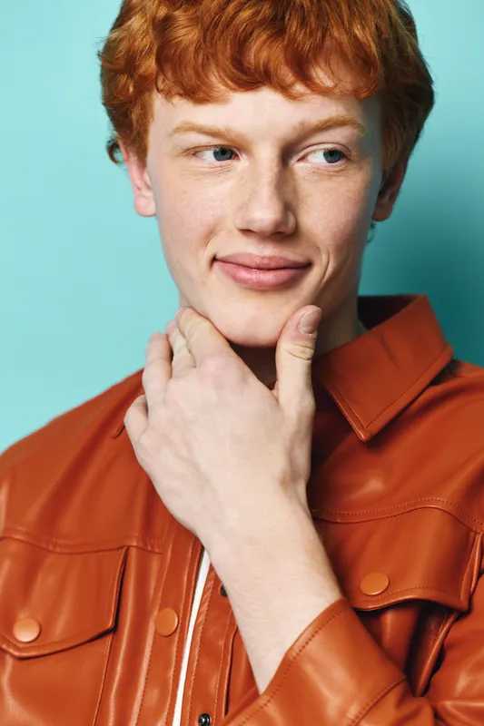 Young redhead man wearing orange jacket with thoughtful expression touching chin against blue background. Portrait, casual style, closeup, male model with natural skin and light freckles.